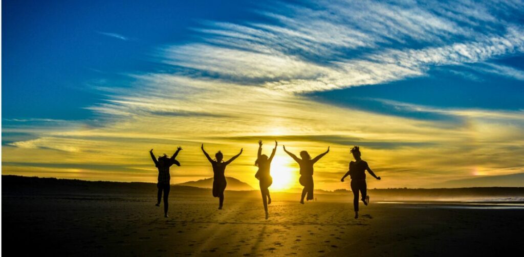 夕暮れのビーチで5人の人々が両腕を上げてジャンプしている逆光のシルエット写真。背景には黄金色とオレンジ色に染まった夕焼け空と青い雲が広がり、太陽が人々の中央に位置している。砂浜には足跡が残り、遠くには海岸線と水平線が見える。喜びと自由を表現した構図。